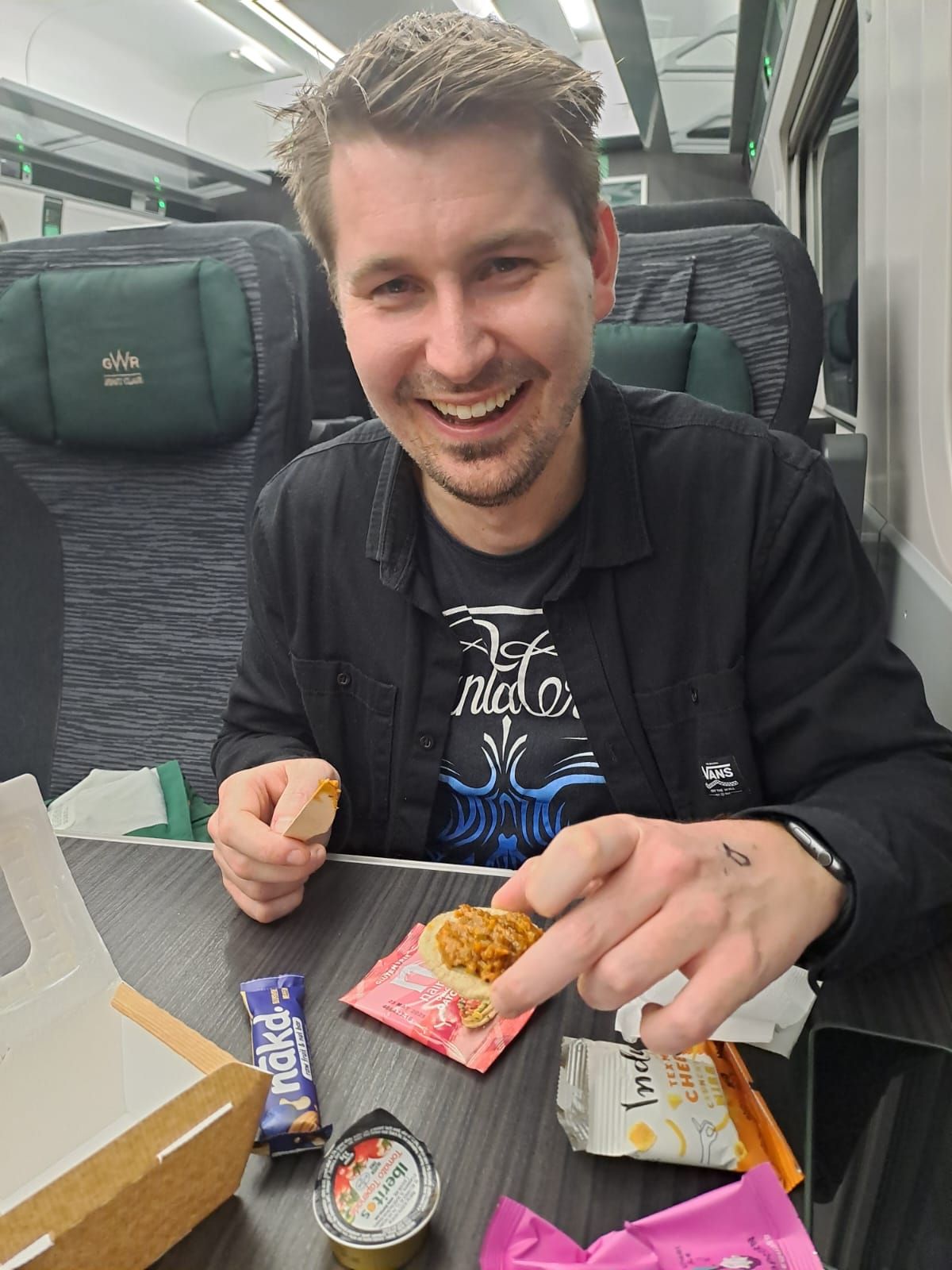 Me smiling ear to ear holding a little cracker covered in tomato tapenade. I'm at a table in a first class GWR train seat. The rest of my vegan selection box is spread across the table in front of me